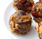 A plate with four round dates laddu sweets, each with a nut topping and a drizzle of syrup on top, arranged on a white background.