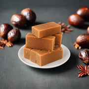 Stack of square-shaped Mysorepak sweets on a white plate with dates and spices around it