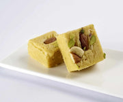 Two pieces of Bannaras Soan papdi, a square-shaped confection made from besan and sugar, garnished with nuts, placed on a white plate.