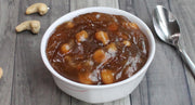 White bowl filled with a brown dessert on a wooden surface with cashews and a spoon.